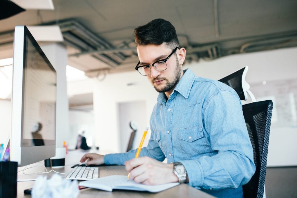 Young attractive dark-haired man in glassess is working with a computer and writing in notebook in office. He wears blue shirt, beard